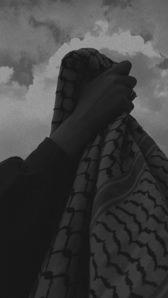 an original keffiyeh held by hand featuring the cloudy sky behind and the black and white pattern.
