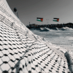 Large Palestinian keffiyeh spread like a landscape with Palestinian flags in the background, used as the cover image for a keffiyeh pronunciation guide.
