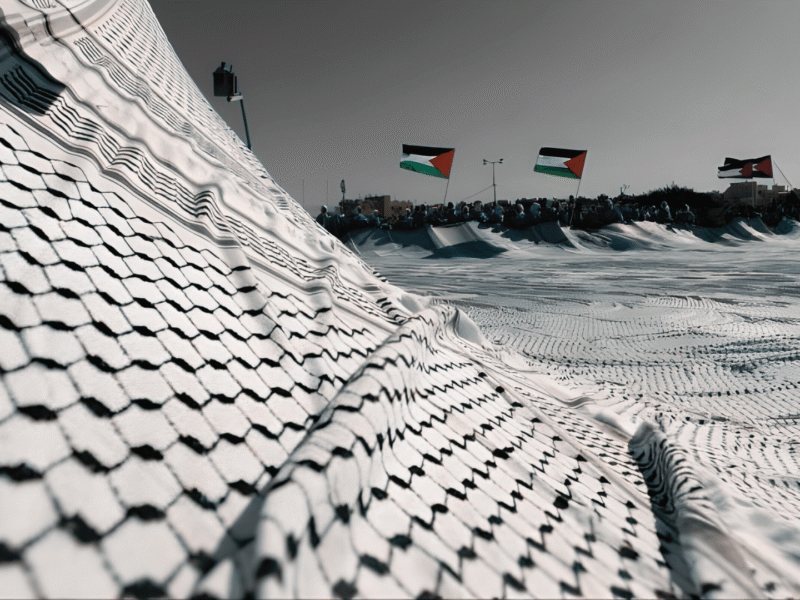 Large Palestinian keffiyeh spread like a landscape with Palestinian flags in the background, used as the cover image for a keffiyeh pronunciation guide.